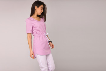 young stylish nurse with long brunette hair in pink medical costume is standing in profile, looking down and smiling cute on the gray wall background. medical fashion concept. free space on right side
