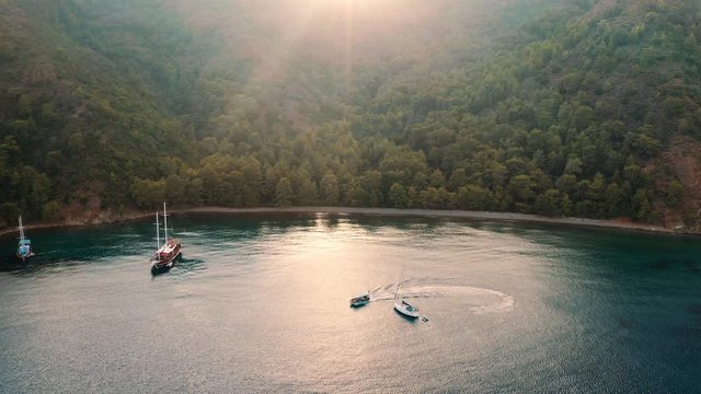 Aerial: Boats park at the private bay with beach, while in the background the sun goes down behind the mountain. Fethiye, Turkey.