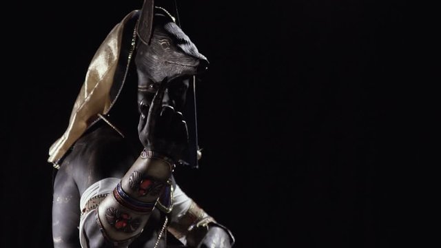 Anubis is sitting in dramatic lighting and beckoning with his hand
