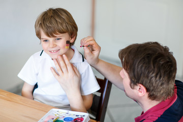 Young dad painting flag on face of little son for football or soccer game. Happy excited preschool...