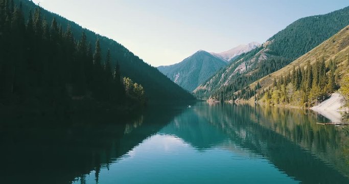 Beautiful landscape of mountain lake. Emerald water surrounded by coniferous forest, autumn grass and passing clouds. Romantic setting, a couple swimming on a white boat. Shooting with, with the drone