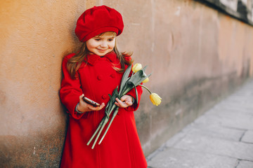 little beautiful girl in red beret and red coat walking in the autumn city with a bouquet of yellow tulips