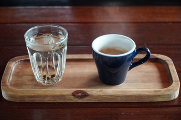 Hot espresso coffee in blue mug with tea glass on wooden tray.