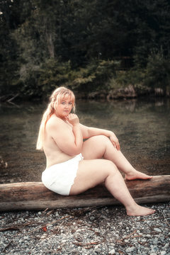 Junge kurvige Frau sitzt wenig bekleidet an einem See auf einem Baumstamm
