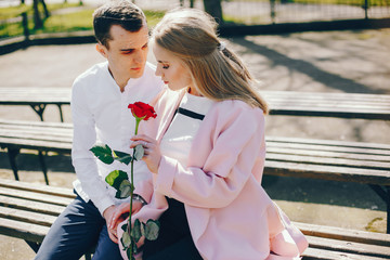 Naklejka premium beautiful and stylish blonde along with her handsome guy sitting on the bench in the sunny summer city with red rose