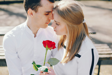 beautiful and stylish blonde along with her handsome guy sitting on the bench in the sunny summer city with red rose