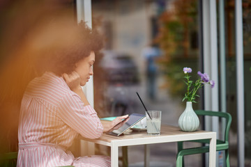 Grogeous mixed race woman in pink striped dress sitting in cafe and using tablet. On table are cup of coffee, book and lemonade.