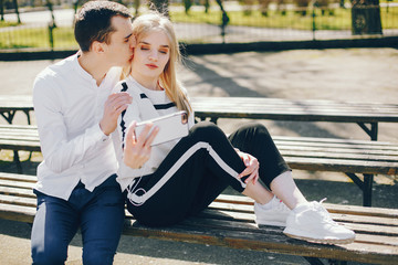 Fototapeta na wymiar beautiful and stylish blonde along with her handsome guy sitting on the bench in the sunny summer city and use the phone