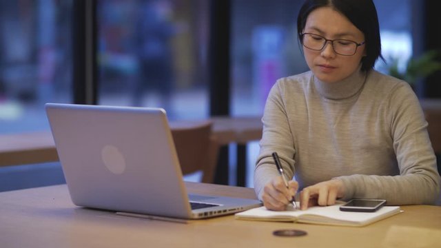 Asian woman studying alone in coffee with phone on table in morning.Chinese woman copying from laptop to notebook search for information, writing down on work-book businesswoman working with laptop 