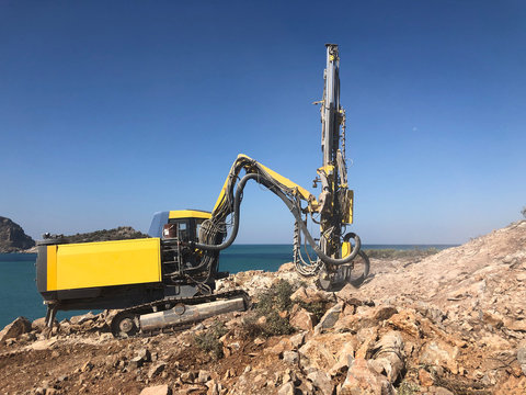 Yellow machinery is drilling holes on the top of rocky sea cliff during blasthole drilling operations