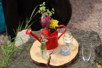 arrangement of a red watering can with flowers inside surrounded by a tea light and flowers on a wooden board