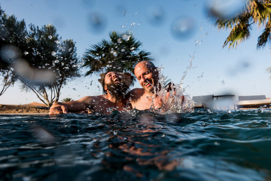 Gay couple in the pool next to palm trees and desert dunes, splashing water.