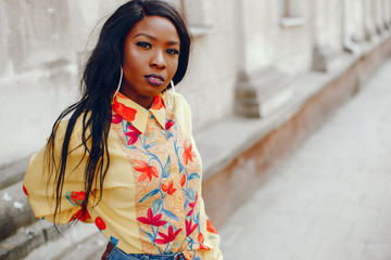 A beautiful and stylish black girl with long dark hair in yellow shirt standing near old building in a summer city