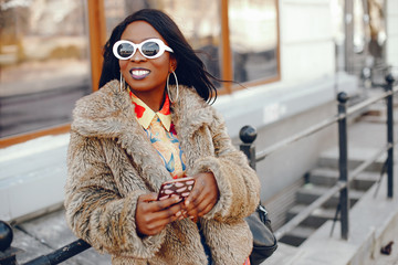 A beautiful and stylish black girl with long dark hair dressed in a brown coat and shirt wallking in a summer city with phone in case