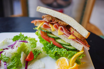 Sandwich with ham cheese baked chicken breast lettuce tomato cucumber salad and orange slices on white plate