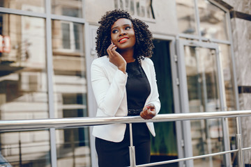 elegant and stylish dark-skinned girl with curly hair and in a white jacket standing in the summer city and using the phone