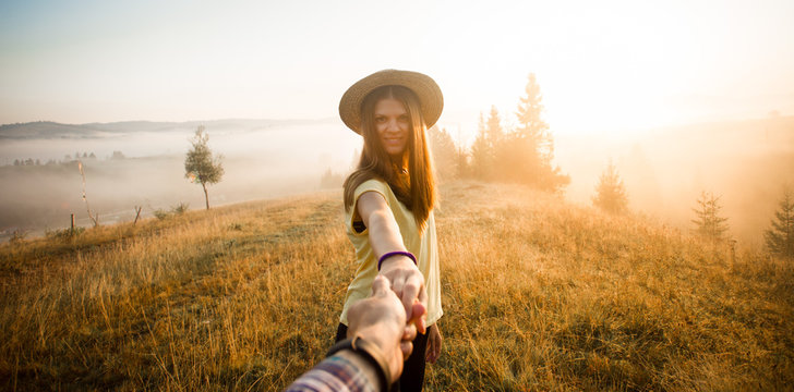Follow me concept. Couple in love holding hands. Woman in yellow shirt and straw hat holding man by hand going to autumn forest with mountains and cloudy sky in morning. Woman leads man.
