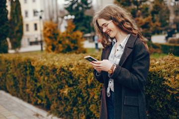 beautiful stylish young girl with long curly hair and a long coat and glasses standing in the autumn city and uses the phone