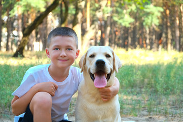 Portrait of cute blond boy hanging out with his pet friend labrador retriever out in the woods. Little guy hugging six months old doggy, walk in a park. Background, copy space, close up.