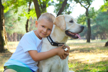 Portrait of cute blond boy hanging out with his pet friend labrador retriever out in the woods. Little guy hugging six months old doggy, walk in a park. Background, copy space, close up.