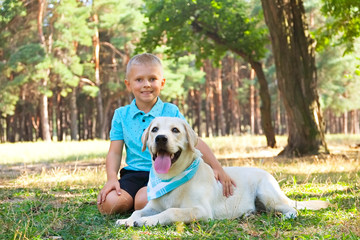 Portrait of cute blond boy hanging out with his pet friend labrador retriever out in the woods. Little guy hugging six months old doggy, walk in a park. Background, copy space, close up.