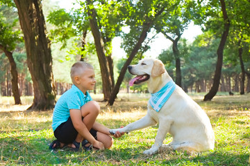 Portrait of cute blond boy hanging out with his pet friend labrador retriever out in the woods. Little guy hugging six months old doggy, walk in a park. Background, copy space, close up.