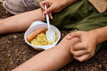 Picnic in the forest. Child's hand holding a plastic spoon in the plate with instant noodles and sausage.