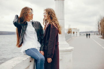 a beautiful stylish young girl with long curly hair and a long coat sitting in the autumn park near river with her girlfriend in a black leather jacket and glasses