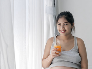 Asian pregnant woman holding a glass of orange juice and standing near window, lifestyle concept.