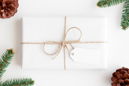 Top View Of A White Christmas Gift With A Blank Tag Surrounded With Pine Cones And Leaves.