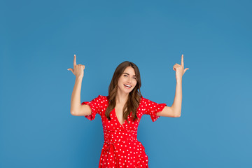 Fototapeta na wymiar Attractive young woman with bright smile in red dress pointing fingers up over blue background.