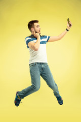 Fototapeta na wymiar Communication. Full length portrait of happy jumping man with gadgets on yellow background. Modern tech, freedom of choices concept, emotions concept. Using smartphone for selfie or videocall in