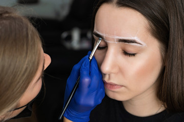 The master makes the procedure for eyebrow correction for a young girl. The girl is dyed with eyebrows with a brush. Facial care in a beauty salon. Marking the shape of eyebrows with a white pencil