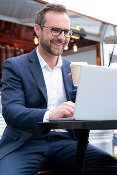 Mature Businessman Working On Laptop By Outdoor Coffee Shop Drinking Takeaway Coffee