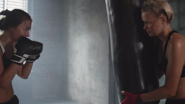 Medium shot of young petty woman wearing sportswear punching heavy bag while female trainer holding it