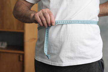 Fat mature man measuring his belly with measurement tape standing in the living room. Problem with overweight, bad health and dieting concept