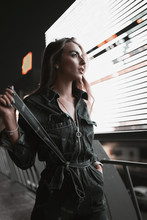 Fashion street portrait of stylish girl in denim overalls on shops background at night. Attractive young woman model walking in city parking in neon light in evening. Urban lifestyle.