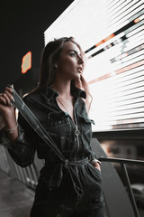 Fashion street portrait of stylish girl in denim overalls on shops background at night. Attractive young woman model walking in city parking in neon light in evening. Urban lifestyle.