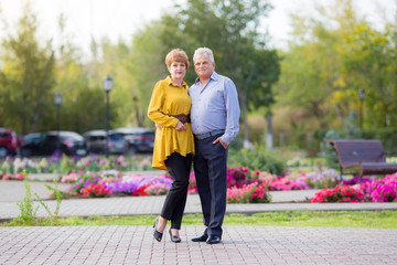 Full-length portrait of a family of senior citizens in a park. A husband and wife of 60 years old are standing next to each other hugging among the flowers.