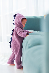 adorable infant baby girl in cute dinosaur costume is standing near the sofa