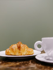 White cup of coffee and saucer and fresh croissant on a wooden table on background of a green olive-green wall for breakfast, lunch, dinner.Vertical