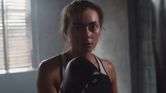 Close up of pretty young woman wearing boxing gloves kicking heavy bag and then looking at camera