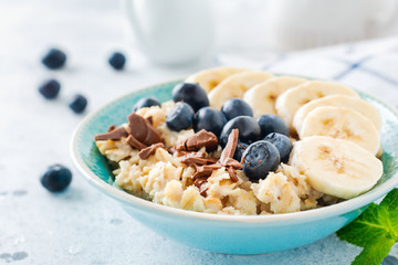 Oat porridge with banana, chocolate and fresh blueberry in a bowl on a light gray slate, stone or concrete background. Top view.