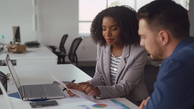 Young smiling african businesswoman explaining new ideas and plans to male colleague holding chart in hand at desk in office