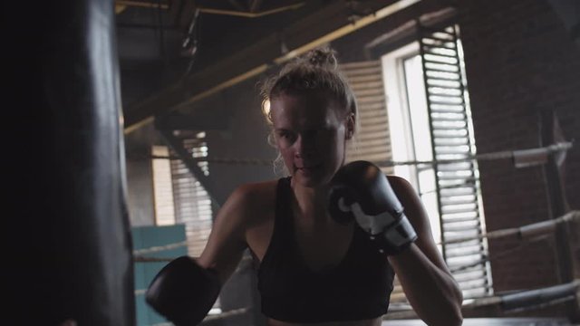Medium shot of slim muscular blonde sportswoman wearing boxing gloves, black racerback tank top and shorts punching boxing bag in gym