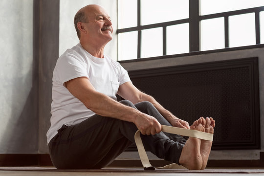 Senior Hispanic Man Sitting In Paschimottanasana Or Intense Dorsal Stretch Pose, Seated Forward Bend Posture, Exercise For Hips And Spine At Home Using Belt.