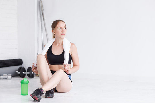 Sporty girl with towel on neck relaxing in gym after hard training, personal trainer after lesson resting sitting on floor with bottle of water, athletic woman taking break before exercises