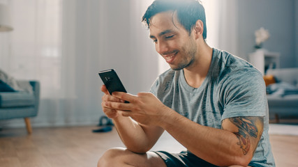 Happy Strong Athletic Fit Man in T-shirt and Shorts is Using a Mobile Phone After Morning Exercises at Home in His Spacious and Bright Living Room with Minimalistic Interior.