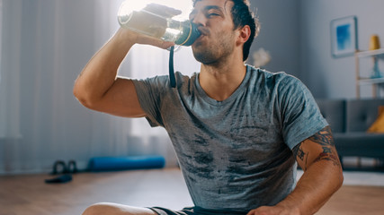 Happy and Satisfied Strong Athletic Fit Man in T-shirt and Shorts is Drinking Water After Morning Exercises at Home in His Spacious and Bright Living Room with Modern Interior.