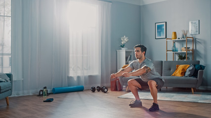 Muscular Athletic Fit Man in T-shirt and Shorts is Doing Squat Exercises at Home in His Spacious...